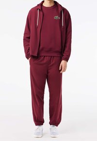Tuta borgogna con giacca con cappuccio a zip e cordini, pantaloni abbinati con polsini elasticizzati e un logo verde sul petto.