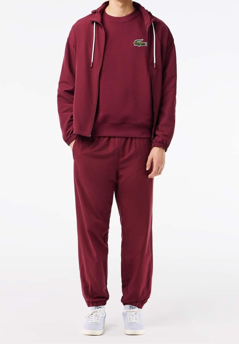 Tuta borgogna con giacca con cappuccio a zip e cordini, pantaloni abbinati con polsini elasticizzati e un logo verde sul petto.