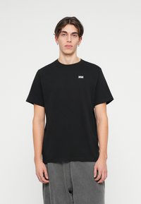 T-shirt de algodão preto com um decote redondo e mangas curtas, apresentando um pequeno logo branco "HH" na área do peito esquerdo. O modelo está a usar calças cinzentas.