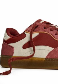 Zapatilla de ante marrón y beige con suela de goma, que presenta costuras detalladas y un cordón marrón desatado que se extiende hacia afuera.