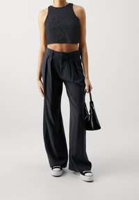 Černý krajkový crop top v kombinaci s černými, vysokým pasem a plisovanými širokými kalhotami a černými teniskami. Malá černá kabelka je držena v ruce.