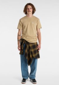 Beige tričko s kapsovým výstřihem a bílým logem, kombinované s volnými modrými džíny a černou a žlutou kostkovanou košilí uvázanou kolem pasu. Černé boty dotvářejí celkový vzhled.