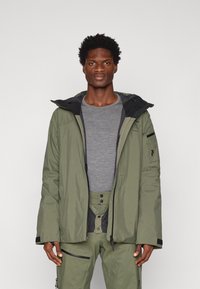 Olíva zöld vízálló dzseki fekete kapucnival, cipzárakkal és zsebekkel. Simább anyag különböző textúrákkal és funkcionális dizájnnal.