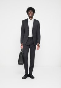 Dunkelgrauer Anzug mit einem taillierten Sakko und schmalen Hosen, einem weißen Hemd darunter, schwarzen Schuhen und einer strukturierten schwarzen Tasche. Glatter, formeller Look.