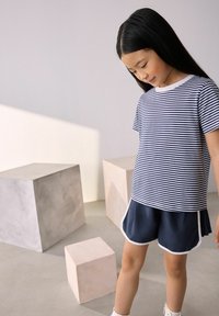 T-shirt rayé bleu marine et blanc à manches courtes associé à un short bleu marine avec des bordures blanches, sur un fond de blocs géométriques abstraits.