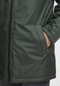 Zielona kurtka ocieplana z zamkiem błyskawicznym, kieszenią boczną oraz zapięciem na napy na mankietach. Gładka faktura materiału, średniej długości design.