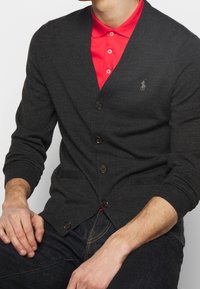 Dunkelgrauer Cardigan mit V-Ausschnitt, Knopfverschluss und zwei vorderen Taschen. Getragen über einem leuchtend roten Poloshirt mit einem kleinen Logodetail.