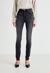 Jeans de mezclilla negra ajustados con cintura alta, con diseño clásico de cinco bolsillos y un desvanecido sutil. Combinados con tacones negros de punta.
