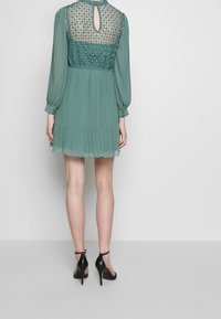 Donna che indossa un abito verde a maniche lunghe, lungo fino al ginocchio, con dettagli in pizzo sulla schiena e gonna plissettata, abbinato a sandali neri con tacco alto.