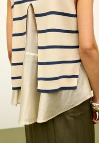 Beżowy sweter z dzianiny w granatowe paski, z rozcięciem z tyłu i warstwą białego materiału pod spodem, w zestawieniu z oliwkowymi spodniami.