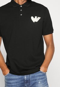 Svart polo skjorta i bomull, med treknappars-öppning, korta ärmar och en krämfärgad hjärta och 'V' logotyp på vänster bröst.