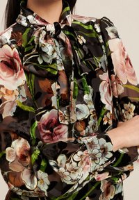 Blusa con estampado floral sobre fondo negro, que presenta flores de múltiples colores, un escote con lazo y mangas largas con puños abotonados.