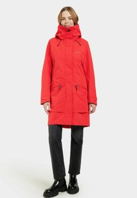 Parka rouge imperméable avec un col haut, une capuche réglable, deux poches latérales, une fermeture éclair devant et une coupe décontractée. Porte sur un pantalon noir et des bottes.