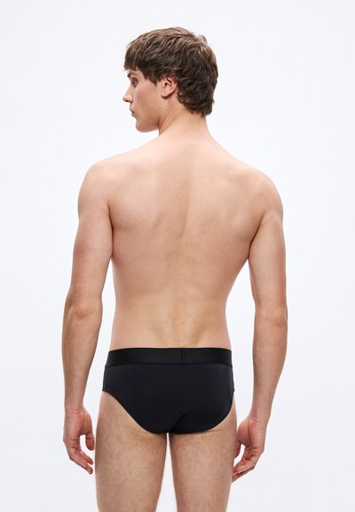 Μαύρο μαγιό τύπου βρακάκι με λεία υφή, ελαστική μέση και μινιμαλιστικό σχέδιο. Φαίνεται από πίσω, μοντέλο σε φυσική στάση.