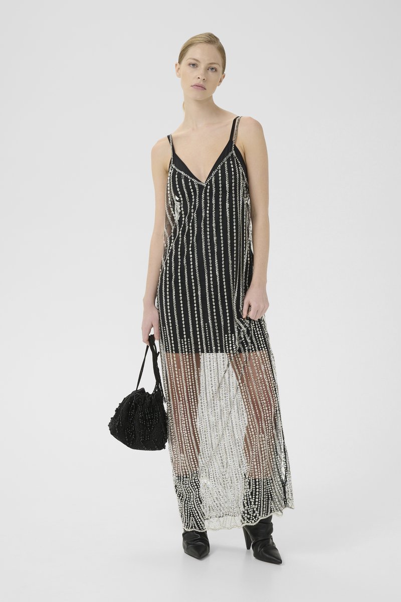 Black and white long dress with vertical bead embellishments, thin straps, deep V-neck, and a sheer overlay. Paired with a textured black bag.