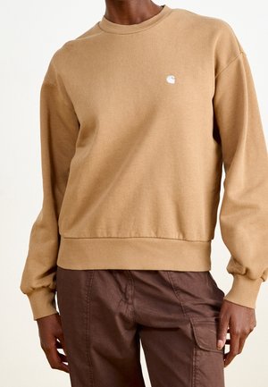 Osoba ubrana w beżową bluzę z nadrukiem bez kaptura, z prążkowanymi mankietami i paskiem u dołu, w połączeniu z ciemnobrązowymi spodniami typu cargo.