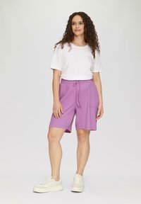 Helle lila Shorts mit einem elastischen Bund und Seitentaschen, kombiniert mit einem weißen T-Shirt mit einem kleinen Druckdesign. Weiße Sneaker.
