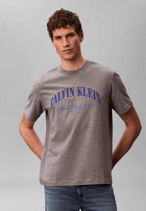 Egy férfi szürke Calvin Klein pólóban, amelyen kék "CALVIN KLEIN New York City" felirat található, háttal a kezét mögé téve, szürke háttér előtt áll.