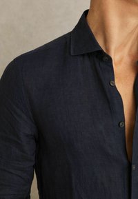 Primer plano de una persona que lleva una camisa azul marino oscura de manga larga con el cuello abierto, mostrando parte del cuello y el pecho.