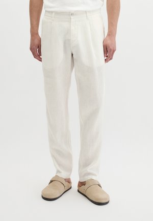 Mannelijk model draagt losse witte linnen broek en beige instapschoenen met zwarte zolen, staand tegen een effen witte achtergrond.