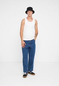 White tank top, blue loose jeans, black bucket hat, and black sneakers with white accents. Model stands with hands in pockets against a plain background.