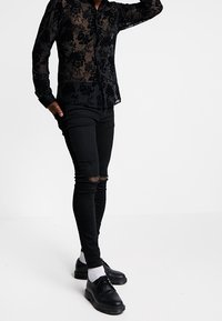 Camicia nera trasparente a fiori con bottoni e maniche lunghe, abbinata a jeans skinny neri strappati e scarpe nere con calze bianche.
