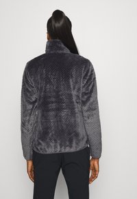 Grå, texturerad fleecejacka med hög krage och mjukt utförande, med ett mönstrat bakstycke och långa ärmar. Inga synliga knäppningar.