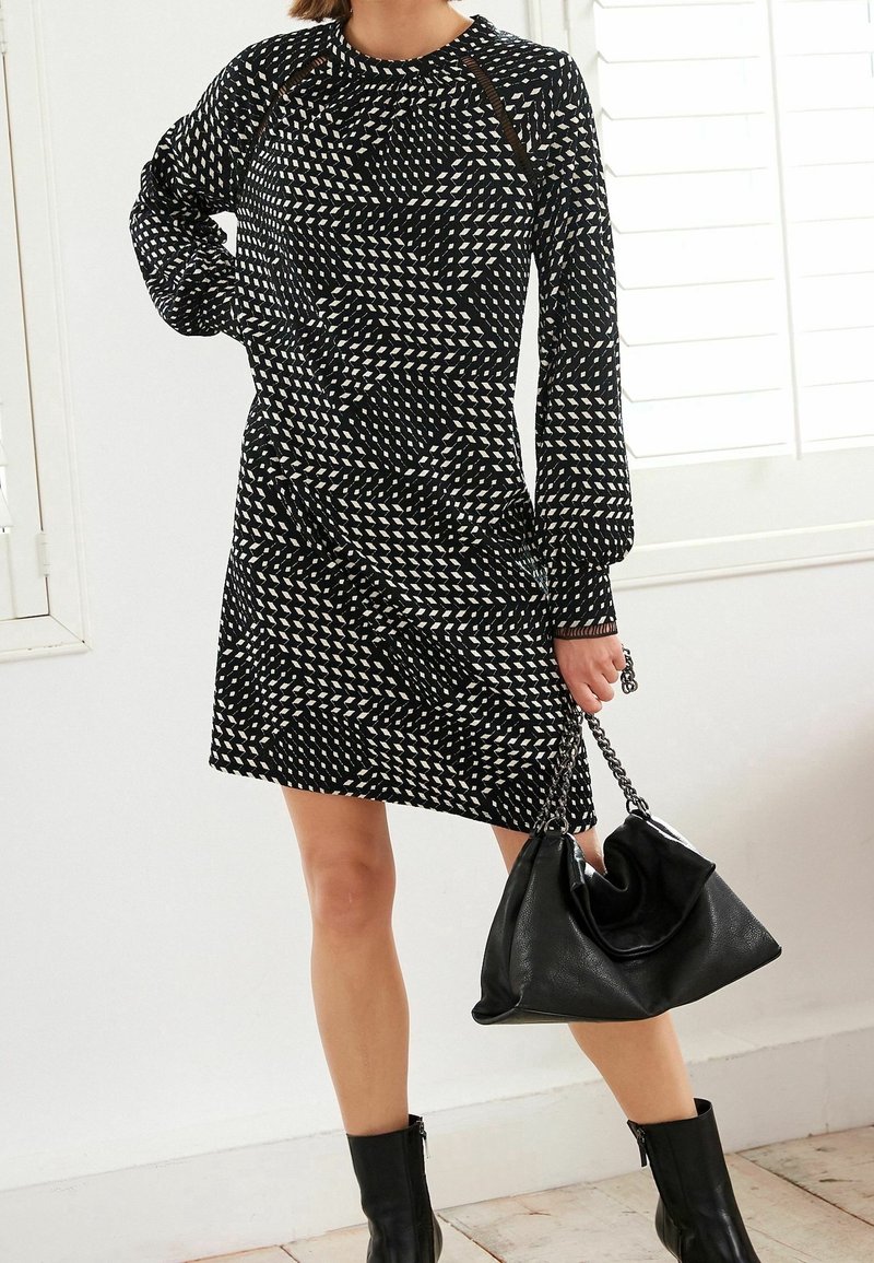 Femme portant une robe à manches longues noire et blanche à motif, des bottines noires, tenant un sac à main noir souple avec des anses en chaîne.