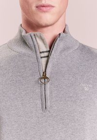 Mann trägt einen grauen Barbour-Reißverschluss-Pullover mit einem Ring-Zipper und darunter eine beige Schicht mit schwarzen Streifen.