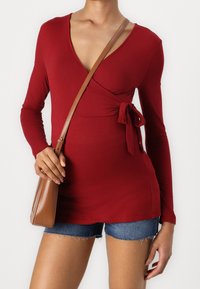 Rotes Langarm-Wickeloberteil mit V-Ausschnitt und seitlichem Bindeband, kombiniert mit Jeansshorts. Eine braune Umhängetasche wird lässig getragen.