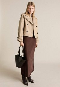 Beige dubbelknäppt jacka med svarta knappar, brun satinkjol, en svart bucket bag och svarta klackstövlar med spännen.