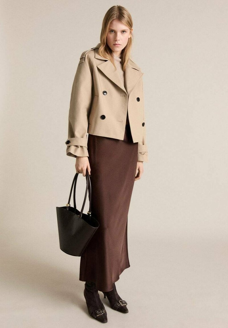 Beige dubbelknäppt jacka med svarta knappar, brun satinkjol, en svart bucket bag och svarta klackstövlar med spännen.