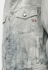 Pelēks džinsa žakete ar teksturētu virsmu, sudraba pogām, krūšu kabatu un nelielu sarkanu logotipa akcentu pie pleca.