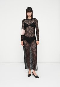 Žena v černých průhledných krajkových šatech s dlouhým rukávem, pod kterými má černou podprsenku a tanga, v černých lodičkách s páskem a s malou světle růžovou kabelkou přes rameno.