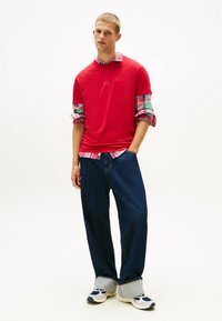 Top rosso a maniche corte con scollatura tonda indossato sopra una camicia a quadri. Pantaloni in denim scuro morbidi e sneakers bianche con dettagli blu.