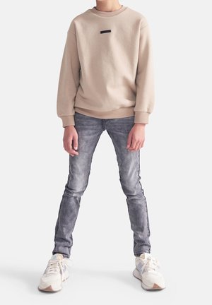 Osoba w beżowym swetrze, poszarpanych szarych dżinsach i białych trampkach stoi z nogami lekko rozstawionymi na białym tle.