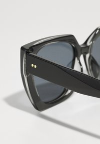 Occhiali da sole neri con montatura spessa e angolare, lenti scure e dettagli dorati sugli steli. Finitura lucida con una texture liscia.