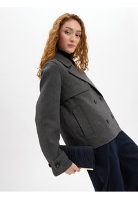 Grijze wollen peacoat met dubbelknopsluiting en een gelapte kraag, gecombineerd met een gestructureerde marineblauwe clutch tegen een witte achtergrond.