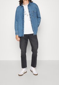 Persona che indossa una camicia di denim blu sopra una maglietta bianca con grafica, jeans neri, calzini bianchi e sneakers bianche con strisce beige.