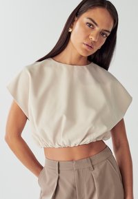 Top corto beige chiaro con scollatura tonda e maniche corte. Presenta un orlo arricciato e una texture di tessuto liscia e leggermente lucida.