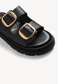 Sandale en cuir noir avec deux larges brides, chacune avec une grande boucle dorée et une semelle épaisse et texturée.