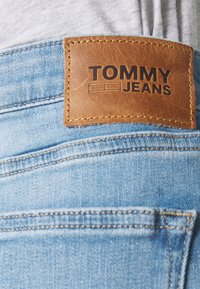 Ljust blå denimjeans har en brun läderlapp med "TOMMY JEANS" präglat. Sömmarna är synliga längs midjebandet och fickorna.