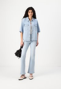 Casaco de ganga azul claro com dois bolsos frontais, combinado com jeans flare azul claro. Os acessórios incluem uma bolsa de cordão preta com padrão floral e sapatos bege.