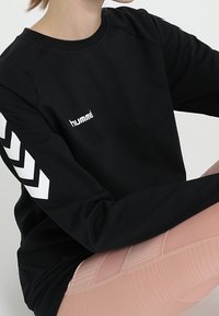 Sweat-shirt noir à manches longues, avec des motifs chevrons blancs sur les manches et un logo sur le devant. Design doux et décontracté.