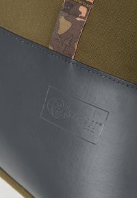 Sac en toile vert olive avec un fond en cuir noir, agrémenté d'un accent en tissu à motif camouflage et d'un logo embossé sur le cuir.