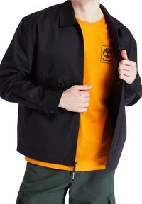 Černá bunda vyrobená z odolné látky, s uzávěrem na zip, hrudní kapsou a límcovým designem, nošená přes oranžovou košili.