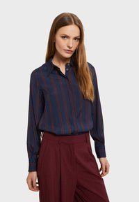 Camicia blu navy con strisce verticali bordeaux, maniche lunghe, colletto a bottoni e texture liscia, abbinata a pantaloni a vita alta bordeaux.