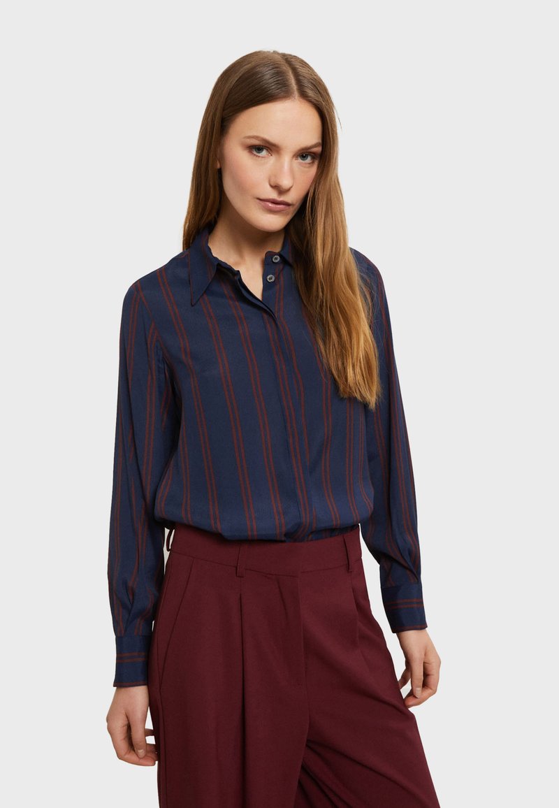 Camicia blu navy con strisce verticali bordeaux, maniche lunghe, colletto a bottoni e texture liscia, abbinata a pantaloni a vita alta bordeaux.