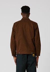 Καφέ καμβάς μπουφάν με γιακά, κουμπωτές μανσέτες και δύο πλευρικά κουμπιά στο στρίφωμα. Υφασμάτινη επιφάνεια με ματ φινίρισμα.