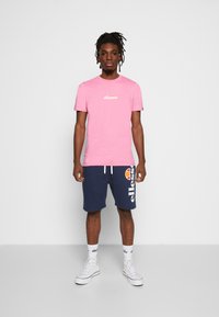 Rosa Kurzarm-T-Shirt mit "ellesse"-Logo, kombiniert mit navyfarbener Shorts mit Logo und runder Akzentuierung, getragen mit weißen High-Top-Sneakern.